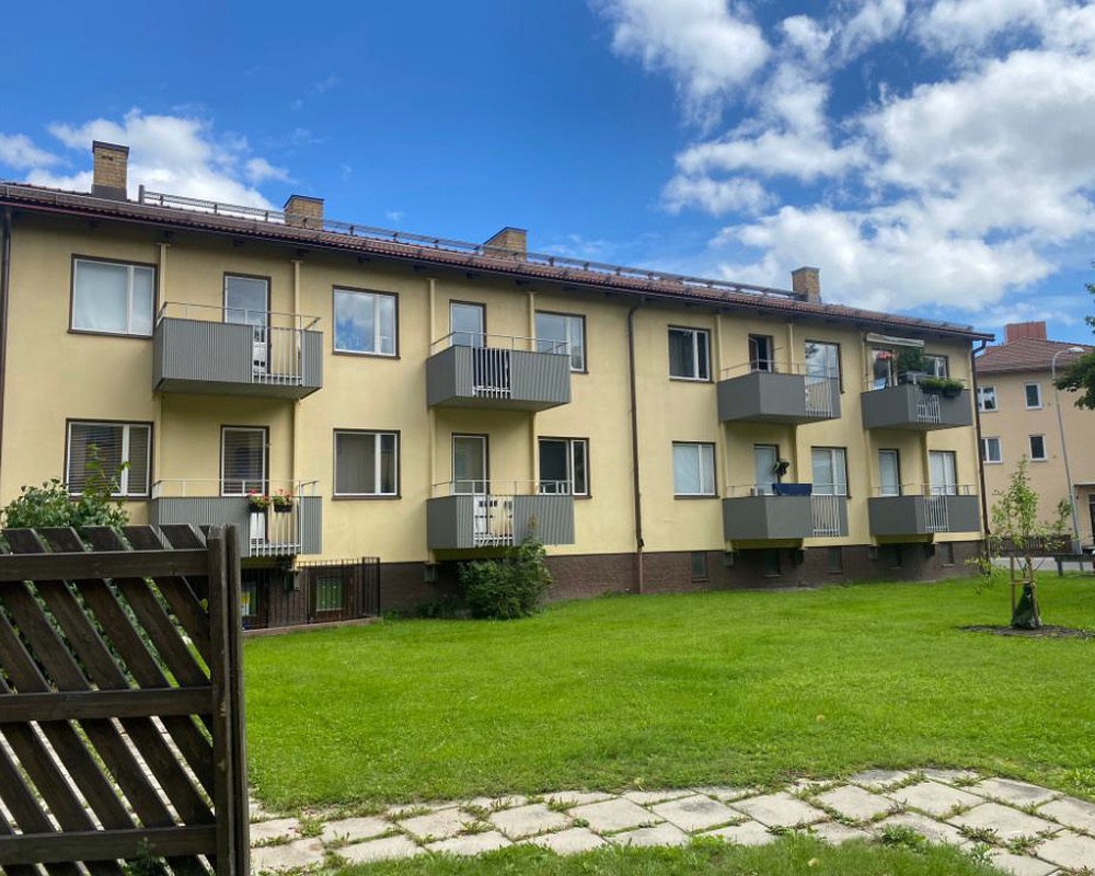 Gult flerbostadshus med grå balkonger, en stor gräsmatta och blå himmel med vita moln