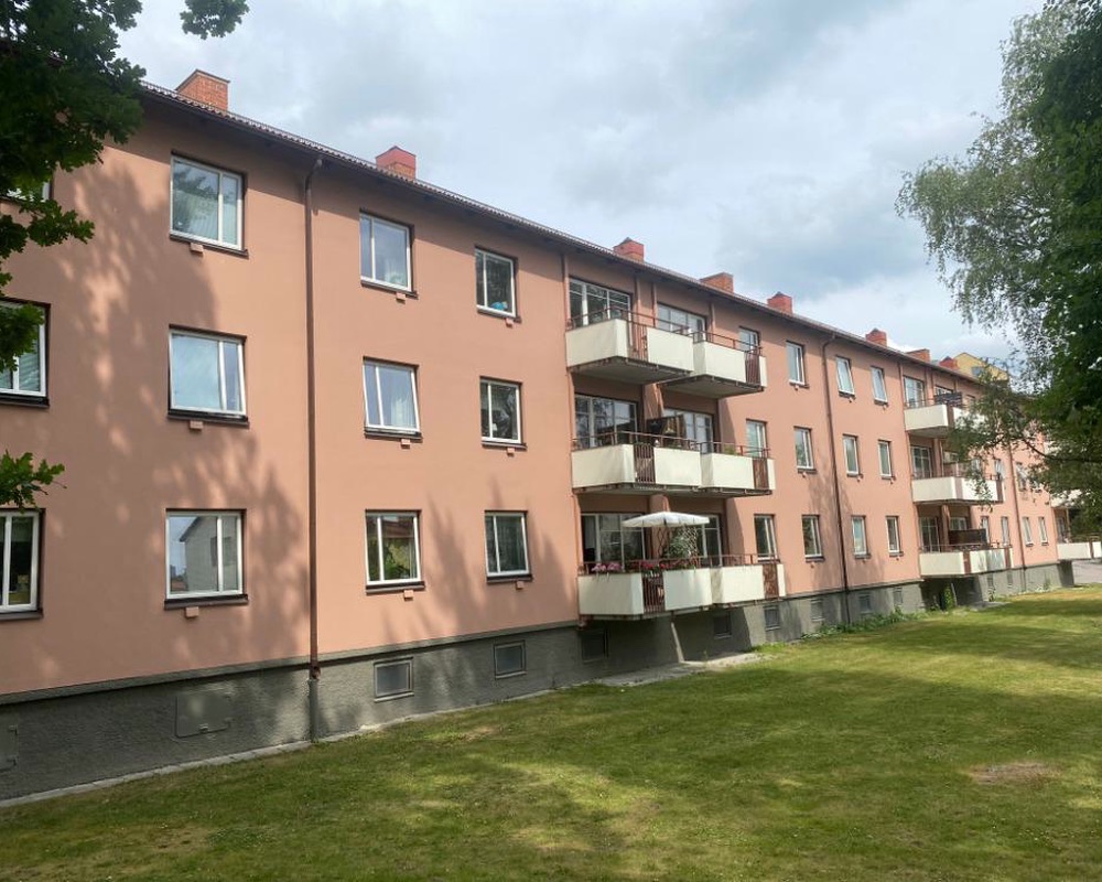 Långt laxrosa flerbostadshus med ljusa balkonger, lövträd och en stor gräsmatta