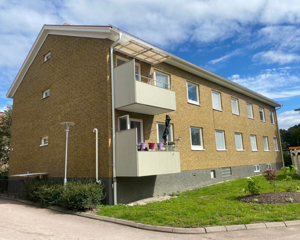 Flerbostadshus i gult tegel med ljusa balkonger, gräsmatta och planteringar under blå himmel med moln