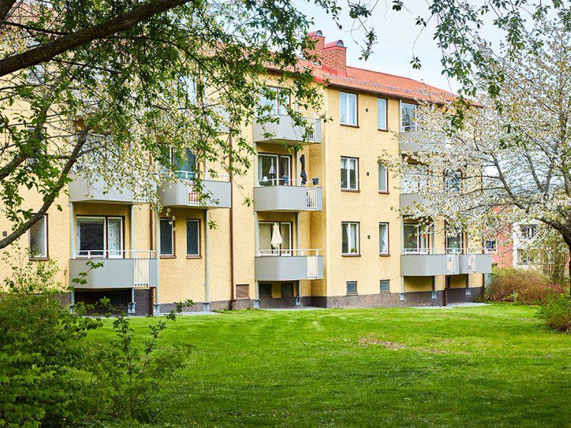 Gult flerbostadshus med balkonger bakom en grön gräsmatta och blommande träd