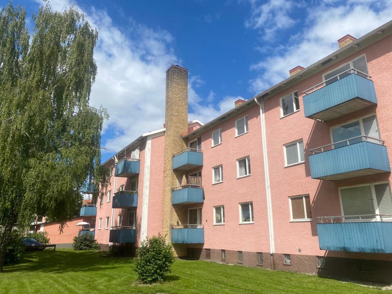 Rosa flerbostadshus med blå balkonger, en hög skorsten, björkar och gräsmatta